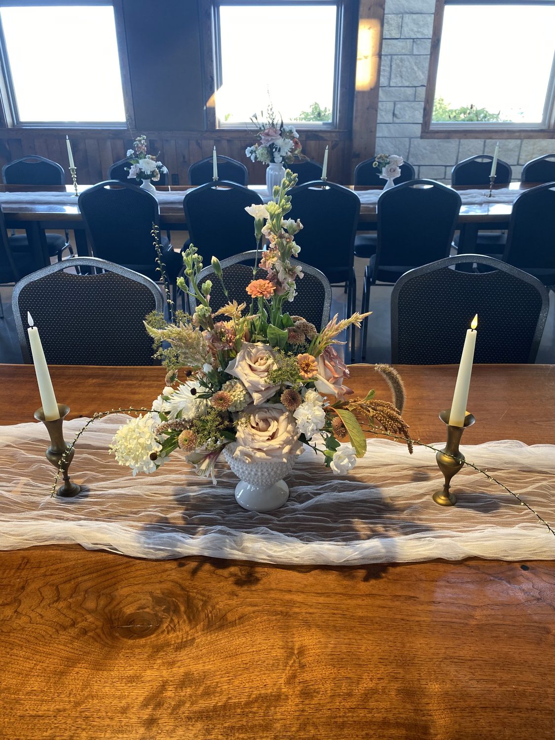 A milk glass centerpiece of toffee roses, zinnias, stock and celosia between brass candlesticks
