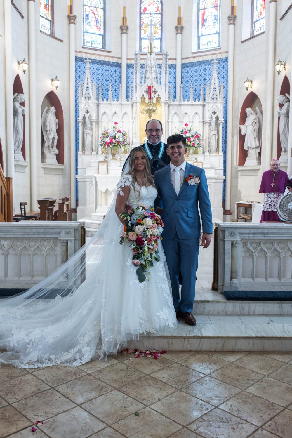 The bride and groom with the priest before the high altar after their Nuptial Mass, her cascade bouquet matched to the sanctuary flowers