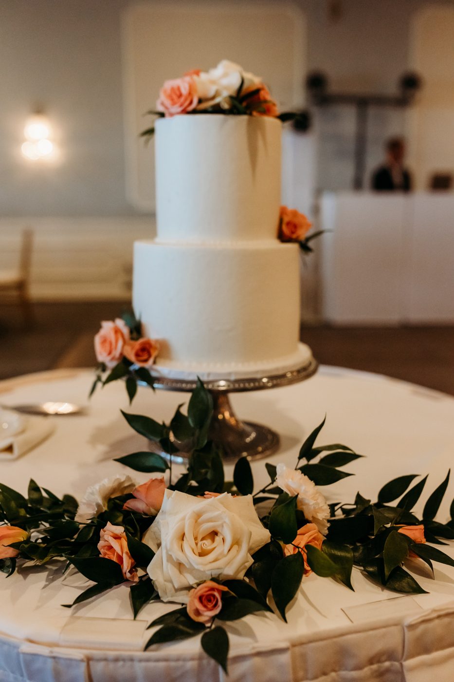 Peach and ivory roses with trailing Italian ruscus on a two-tier buttercream cake, with a matching garland around the pedestal