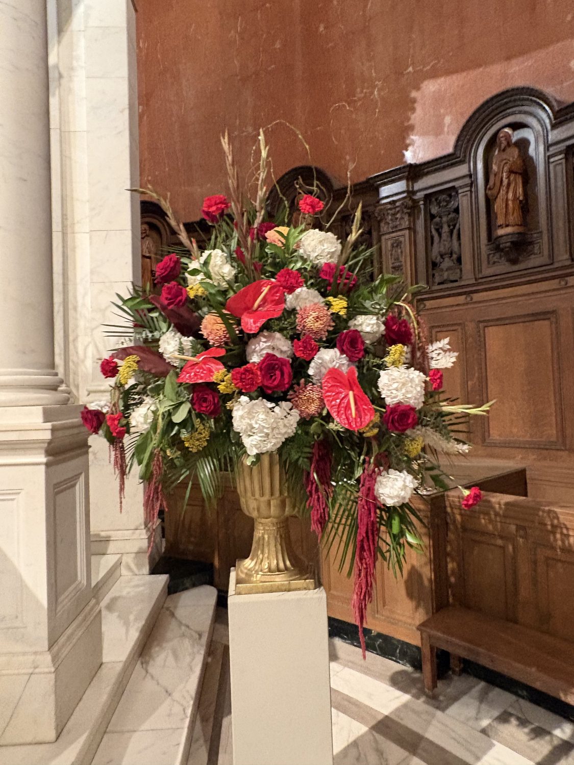 A large gold urn arrangement of red anthurium, roses, white hydrangea and trailing amaranthus on a pedestal in the sanctuary