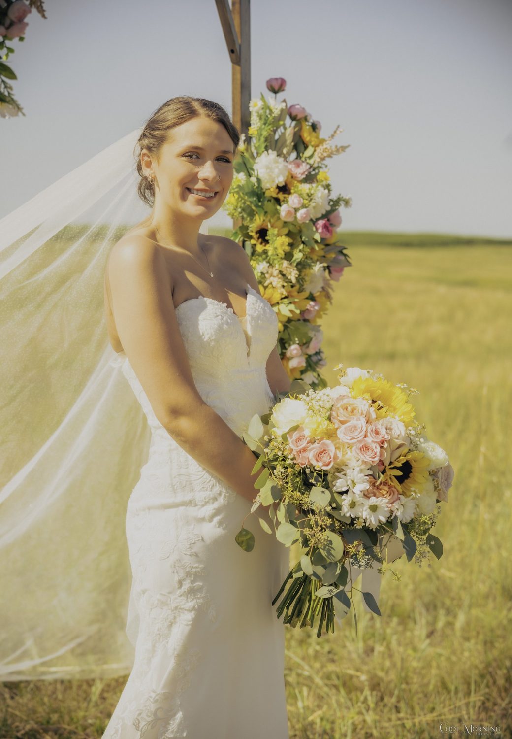 The bride beside the arch’s cascade of sunflowers and pink roses