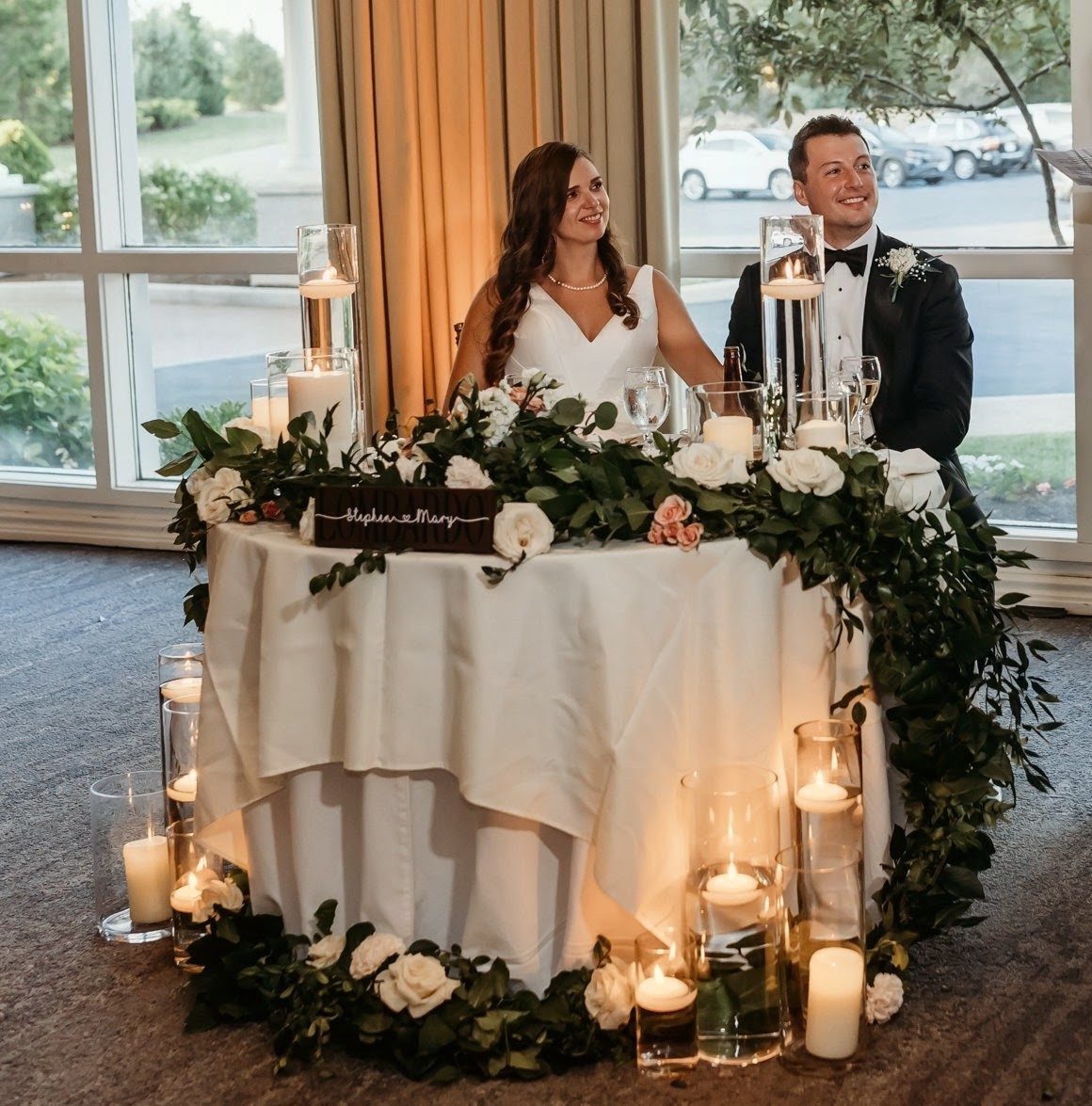 A sweetheart table draped in white roses, blush spray roses and cascading greenery, glowing with pillar and floating candles